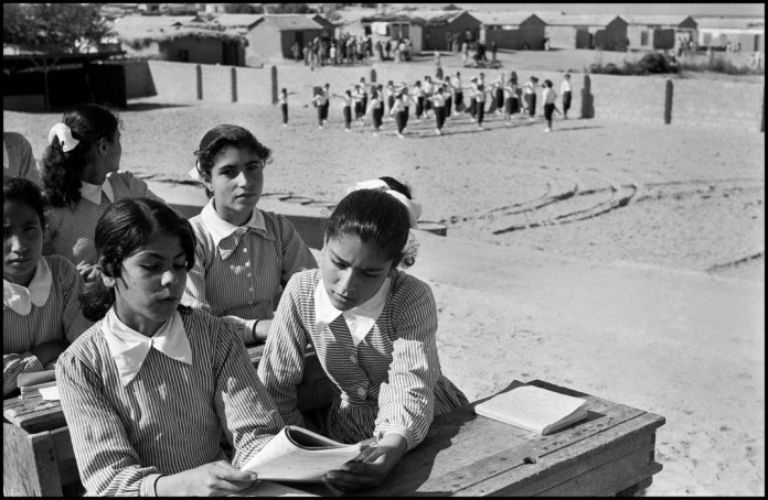DEIR EL BALAH (The Monastery at the Sea) Camp in Jabalya near Gaza.This is one of the best camp schools for refugee girls.They wear self made uniforms, desks have been made by refugee boys in the carpentry shop. In the background girls are having a sports class, behind the schoolyard, are the mud huts of the camp.